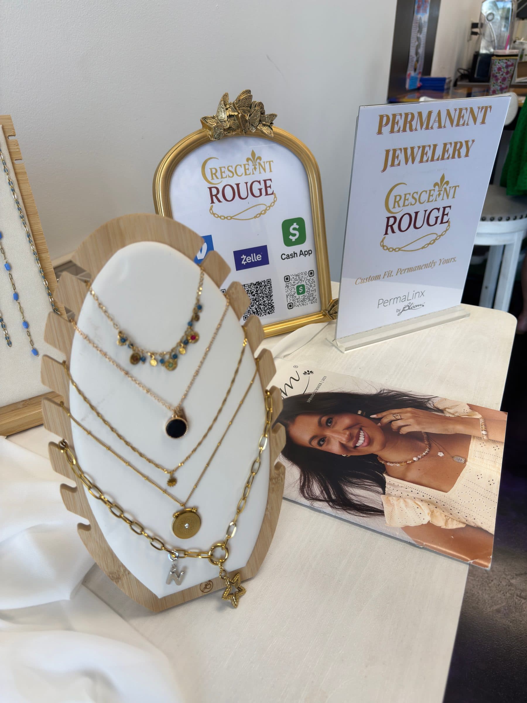 Gold necklaces on a white display beside the Crescent Rouge permanent jewelry signs